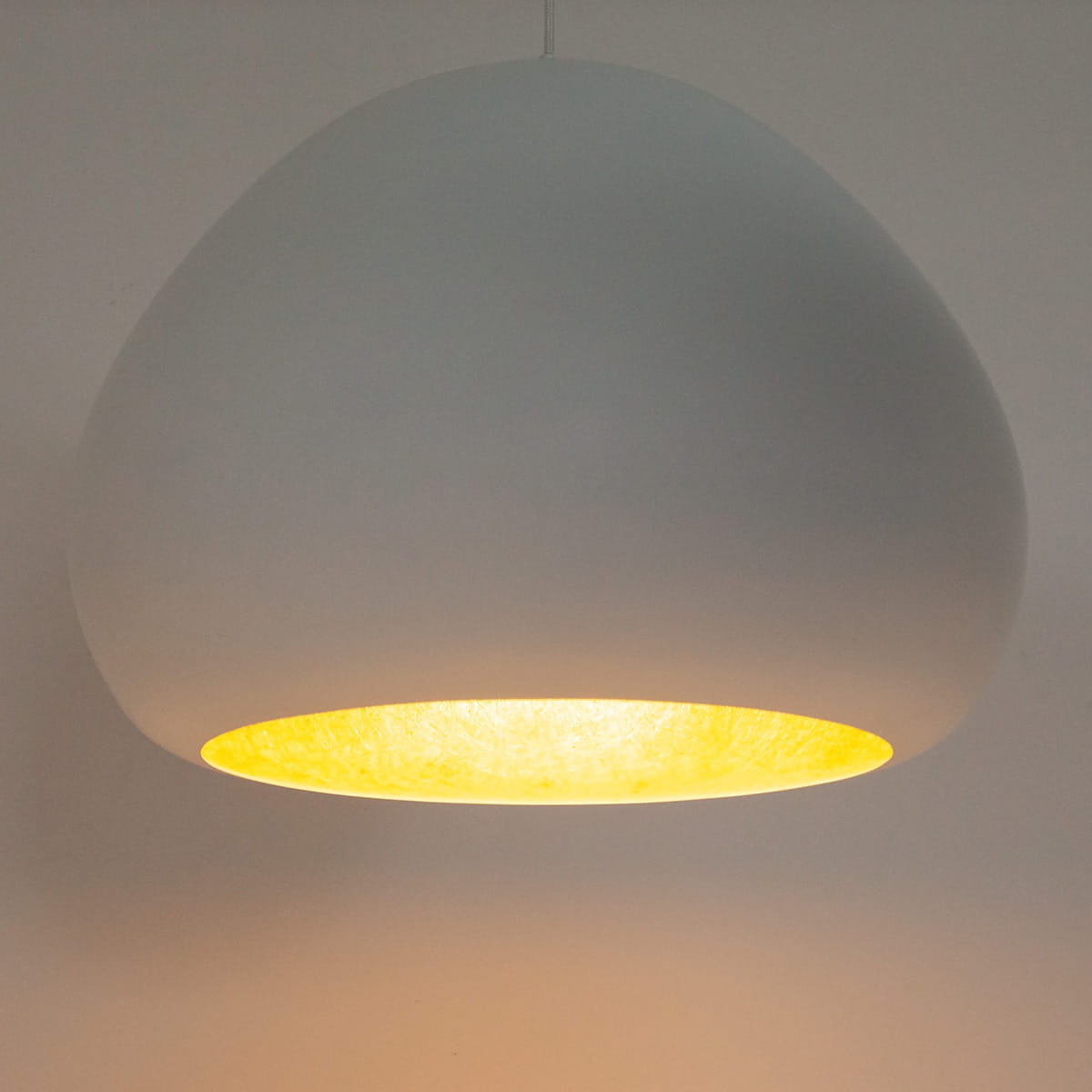 Biała lampa wisząca Lava D52 z kloszem w kształcie owalnym, z wewnętrzną złotą powłoką, zawieszona na cienkim przewodzie. Widoczny efekt ciepłego światła emitowanego z wnętrza klosza.
