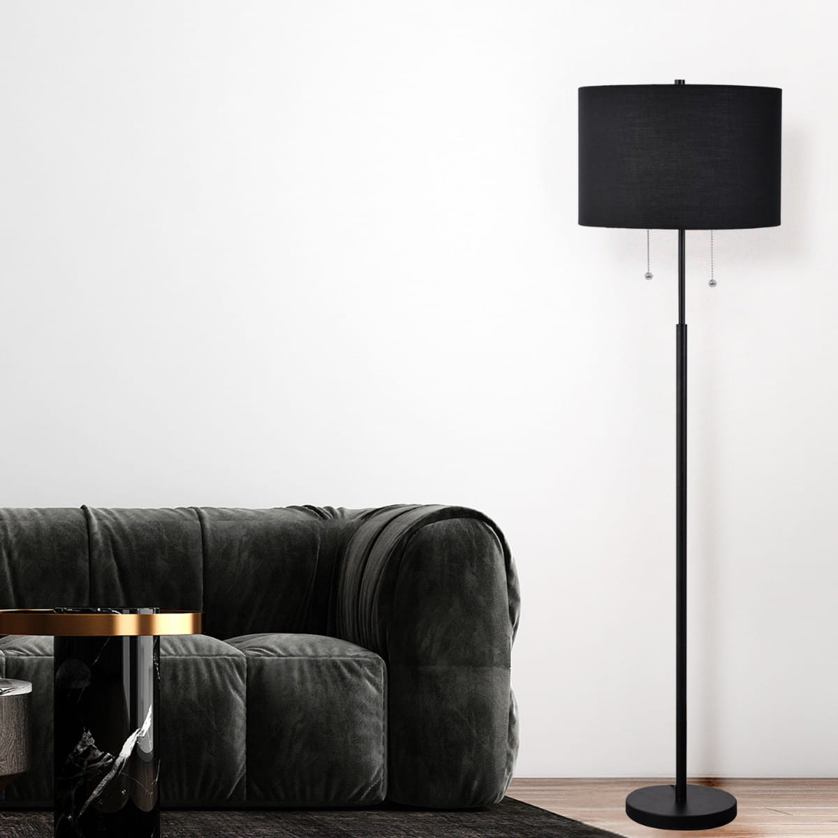 Lampa podłogowa Fogo w kolorze czarnym z kloszem o cylindrycznym kształcie, wyposażona w dwa gniazda E27. Stojąca na okrągłej podstawie, z dwoma metalowymi suwakami do regulacji światła. Ustawiona obok szarej sofy na tle białej ściany.