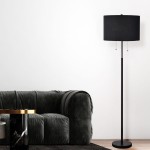 Lampa podłogowa Fogo w kolorze czarnym z kloszem o cylindrycznym kształcie, wyposażona w dwa gniazda E27. Stojąca na okrągłej podstawie, z dwoma metalowymi suwakami do regulacji światła. Ustawiona obok szarej sofy na tle białej ściany.