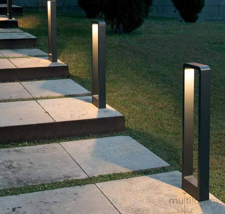 Lampa stojąca ogrodowa Rio LED H60 w antracytowym kolorze, w kształcie prostokątnego słupka, umieszczona wzdłuż ścieżki z płyt kamiennych, emitująca ciepłe światło, otoczona trawnikiem i niską roślinnością.
