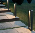 Lampa stojąca ogrodowa Rio LED H60 w antracytowym kolorze, w kształcie prostokątnego słupka, umieszczona wzdłuż ścieżki z płyt kamiennych, emitująca ciepłe światło, otoczona trawnikiem i niską roślinnością.