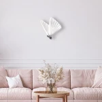 Kinkiet Mariposa 1xLED/8W w kolorze chromu, z dwoma skrzydłami w kształcie liści, zamontowany na jasnej ścianie. W tle widoczna różowa sofa z poduszką i dekoracyjnym stolikiem oraz wazonem z suchymi kwiatami.