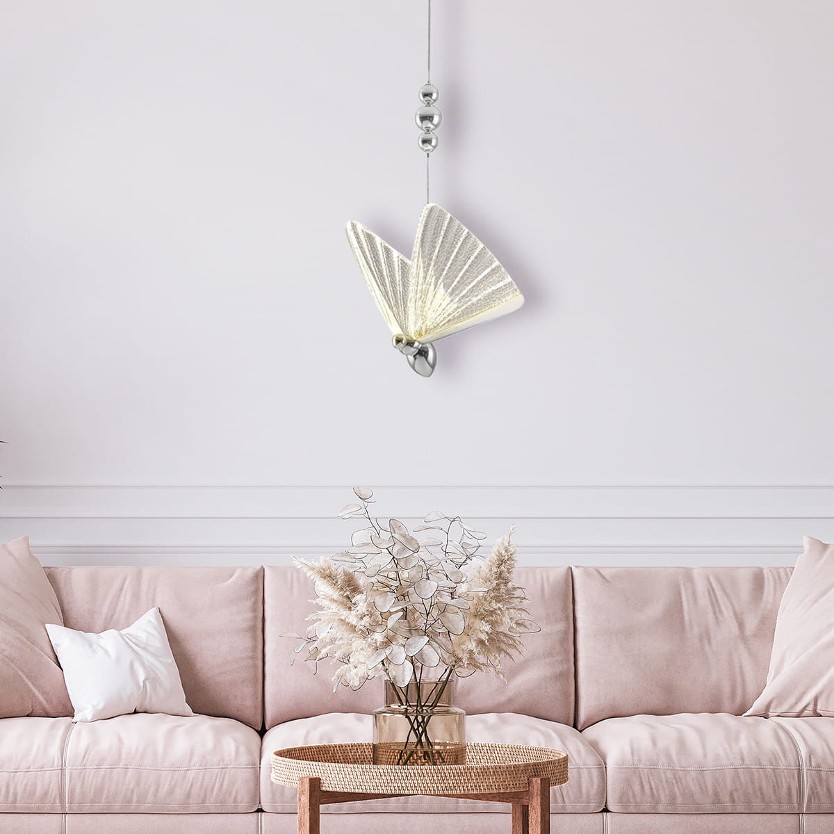 Lampa wisząca Mariposa w kształcie motyla, chromowana, z jedną żarówką LED o mocy 8W. Zawieszona nad różową sofą, w otoczeniu dekoracji roślinnych. Kąt ujęcia frontalny, na jasnym tle.