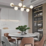 Lampa wisząca Bolti 7xE14 w złotym wykończeniu, z sześcioma białymi, okrągłymi kloszami, zawieszona nad okrągłym stołem w nowoczesnej kuchni. Tło z jasnymi szafkami i marmurowym blatem.