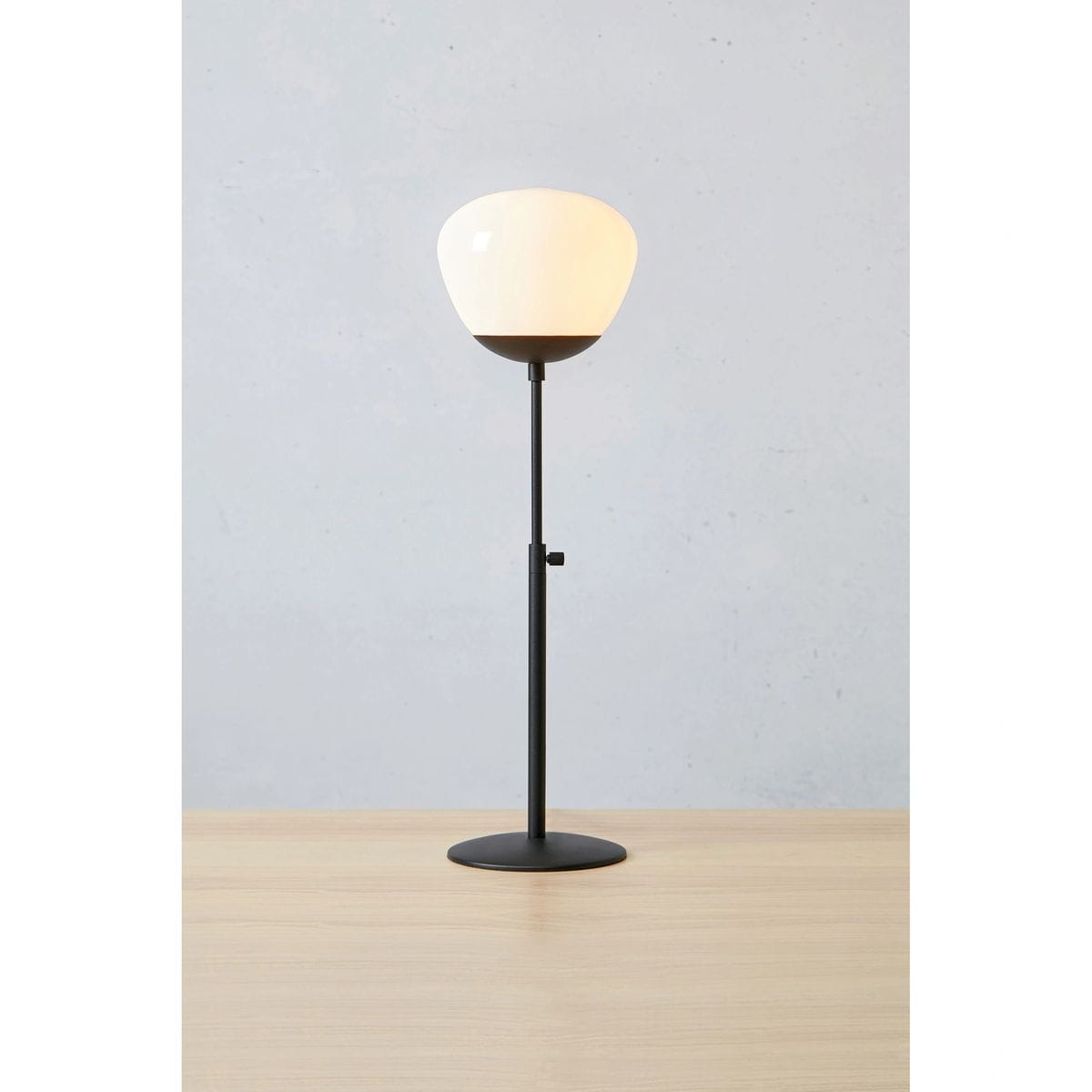 Lampa stołowa Rise H60 z czarną podstawą i białym kloszem o kształcie miski. Ustawiona na drewnianym stole, z regulowaną wysokością. Klosz wykonany z przezroczystego materiału, montowany na cienkim metalowym stelażu.
