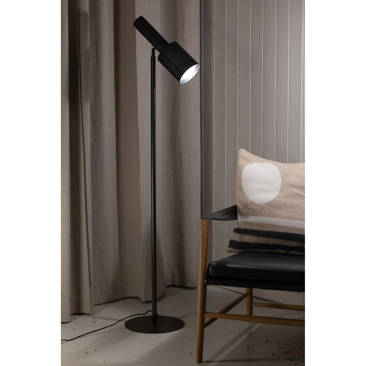 Lampa podłogowa Ozzy w kolorze czarnym, z metalowym kloszem i regulowanym ramieniem. Stoi na okrągłej podstawie, obok czarnego fotela z poduszką w jasnych odcieniach. Tło stanowią jasne zasłony i drewniana ściana.