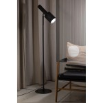 Lampa podłogowa Ozzy w kolorze czarnym, z metalowym kloszem i regulowanym ramieniem. Stoi na okrągłej podstawie, obok czarnego fotela z poduszką w jasnych odcieniach. Tło stanowią jasne zasłony i drewniana ściana.