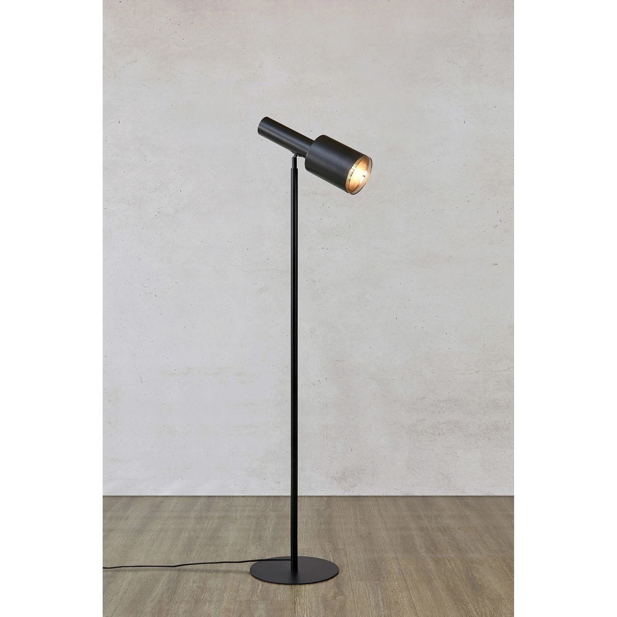Czarna lampa podłogowa Ozzy z jedną oprawą E27, z prostym, cylindrycznym kloszem i długą, smukłą nogą. Ujęcie frontalne, lampa stoi na okrągłej podstawie, w tle jednolita ściana i podłoga w jasnym odcieniu.