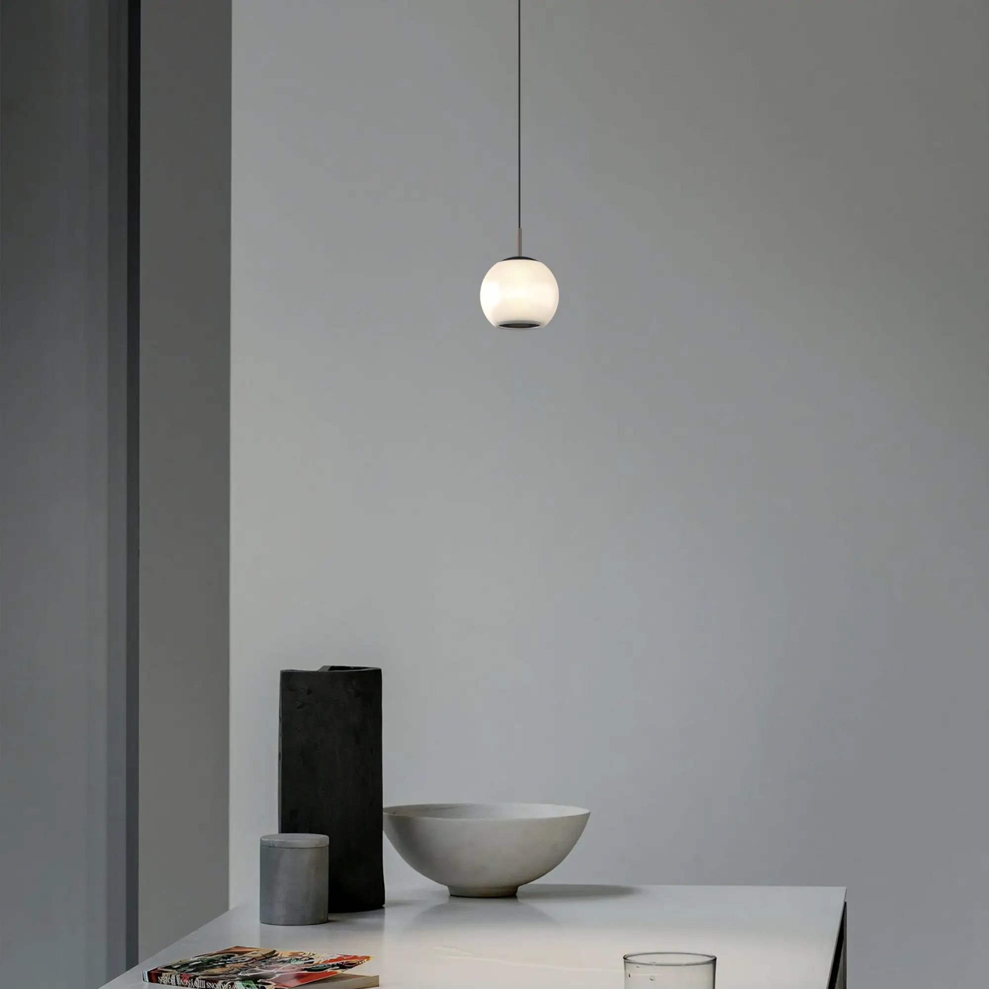 Lampa wisząca Iliso w kolorze czarnym, z okrągłym, białym kloszem, zawieszona na cienkim kablu. Produkt znajduje się nad białym stołem, obok szarej miski i geometrycznych dekoracji. Tło w odcieniach szarości, minimalistyczne otoczenie.