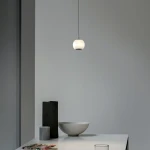 Lampa wisząca Iliso w kolorze czarnym, z okrągłym, białym kloszem, zawieszona na cienkim kablu. Produkt znajduje się nad białym stołem, obok szarej miski i geometrycznych dekoracji. Tło w odcieniach szarości, minimalistyczne otoczenie.