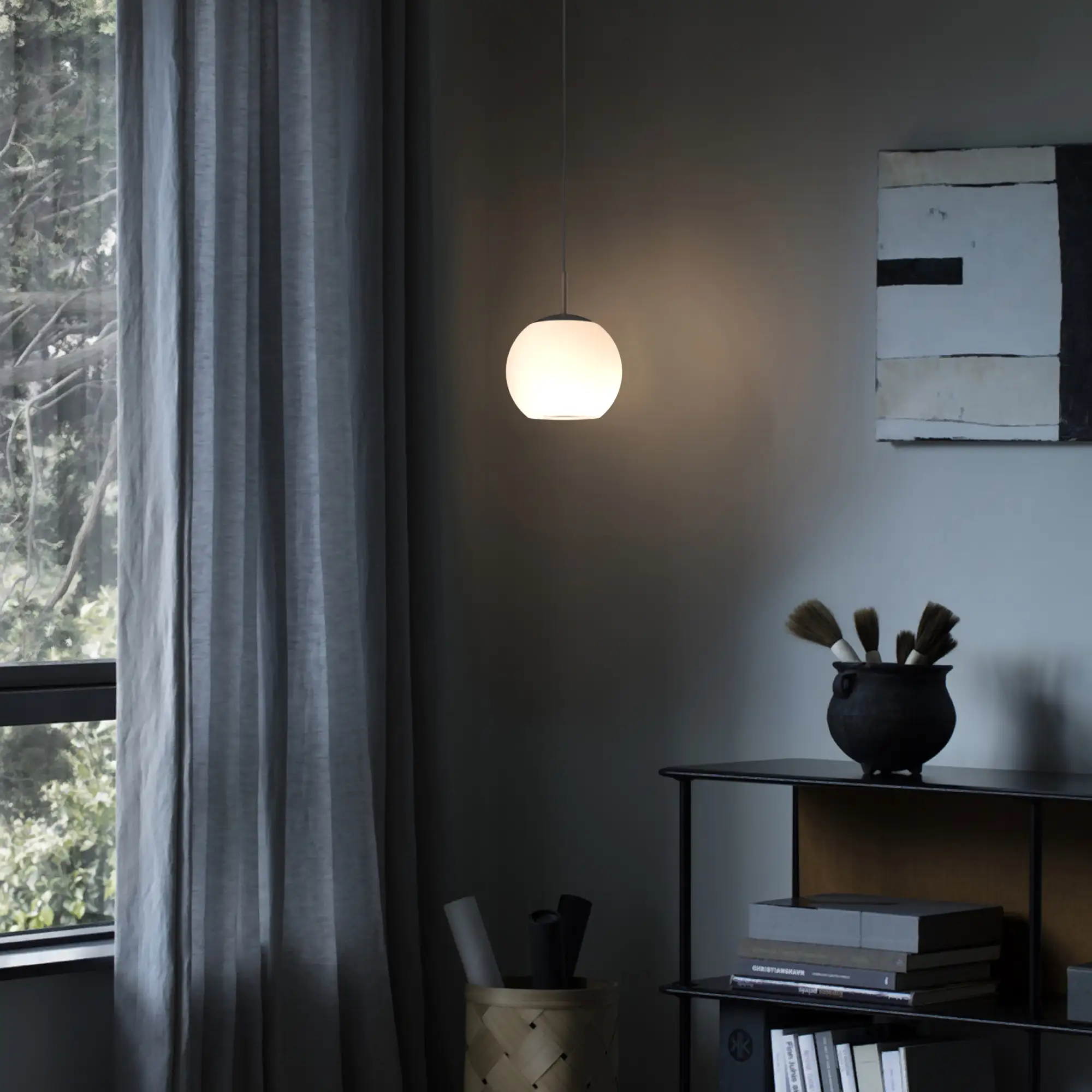 Lampa wisząca Iliso w kolorze czarnym, z okrągłym kloszem z białego szkła, zawieszona na cienkim kablu. Umieszczona w nowoczesnym wnętrzu, obok ciemnej półki z dekoracjami, w tle zasłony i okno. Idealna do oświetlenia przestrzeni.