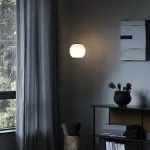 Lampa wisząca Iliso w kolorze czarnym, z okrągłym kloszem z białego szkła, zawieszona na cienkim kablu. Umieszczona w nowoczesnym wnętrzu, obok ciemnej półki z dekoracjami, w tle zasłony i okno. Idealna do oświetlenia przestrzeni.