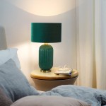 Lampa stołowa w butelkowej zieleni z żłobionym kloszem i metalową podstawą. Ustawiona na drewnianym stoliku obok łóżka, w otoczeniu poduszek i książek. W tle widoczne zasłony.