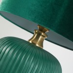 Lampa stołowa w butelkowej zieleni z żłobionym, ceramicznym korpusem i złotym uchwytem. Posiada welurowy klosz w tym samym odcieniu, ustawiona w pozycji stojącej, z bliska ukazuje detale wykończenia.