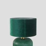 Lampa stołowa w butelkowej zieleni z okrągłym, welurowym abażurem i złotym detalem. Kształt podstawy jest zaokrąglony z pionowymi żłobieniami. Produkt widoczny z boku, na jednolitym tle.
