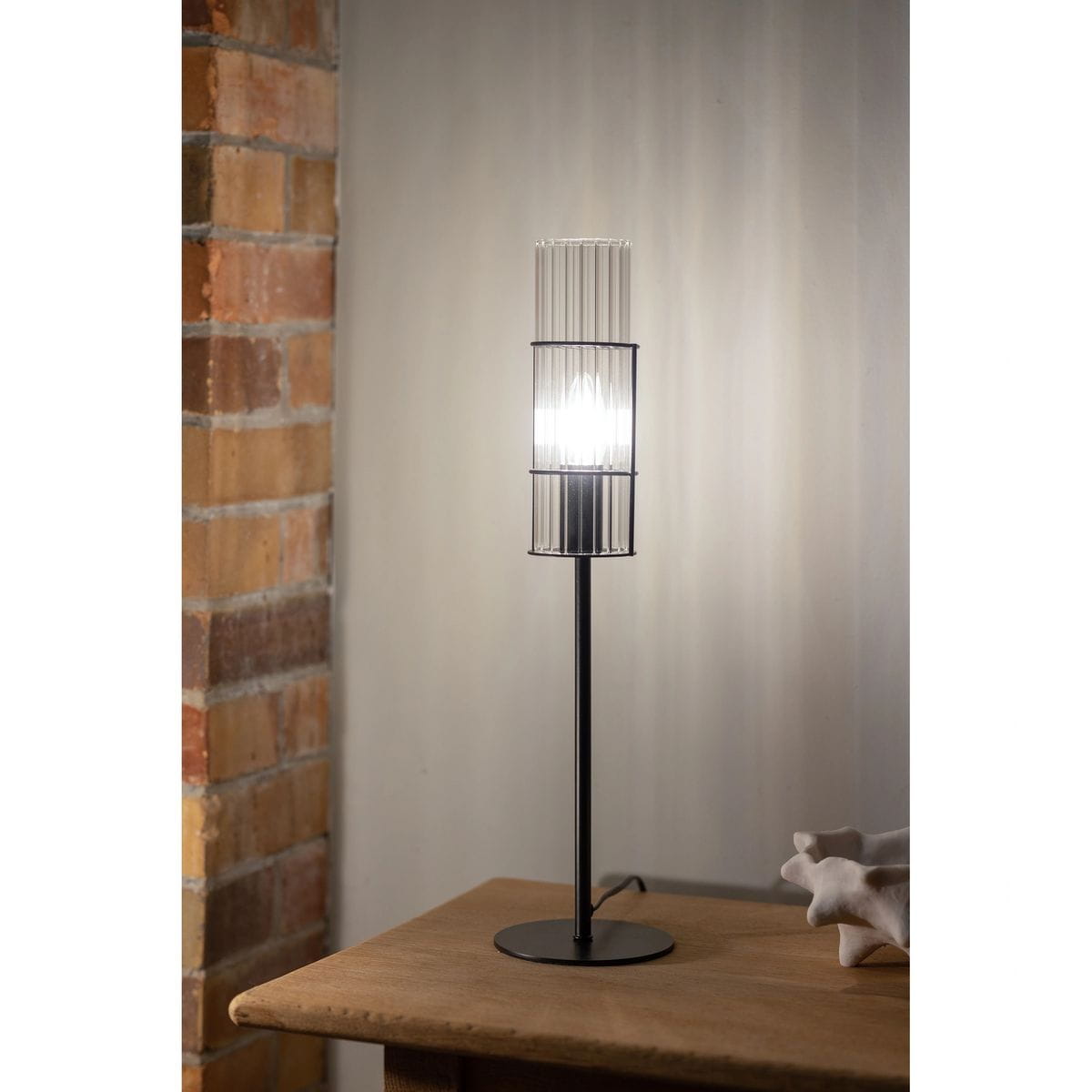 Lampa stołowa Tubo H50 z jednym gniazdem E14, czarna podstawa i przeźroczysta klosz z pionowymi żeberkami. Stoi na drewnianym blacie, w tle widoczna ceglana ściana. Światło emitowane z wnętrza klosza.