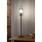 Lampa stołowa Tubo H65, z metalowym czarnym stelażem i przezroczystym kloszem z żebrowanej szkła. Ujęcie z boku, ukazujące źródło światła E14. Stoi na drewnianym stole, w tle ceglana ściana.