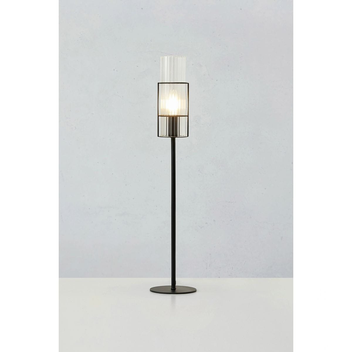 Lampa stołowa Tubo H65 z czarną podstawą i przezroczystym kloszem, stojąca na okrągłej podstawie. Klosz o cylindrycznym kształcie z pionowymi żłobieniami, zawiera miejsce na żarówkę E14. Produkt na białym tle, prezentowany frontalnie.