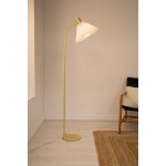 Lampa podłogowa Mira z kloszem w kształcie stożka, wykonana ze szczotkowanego mosiądzu i białego tworzywa. Ujęcie z boku, stojąca na okrągłej podstawie. W tle widoczny fragment krzesła i dywan.