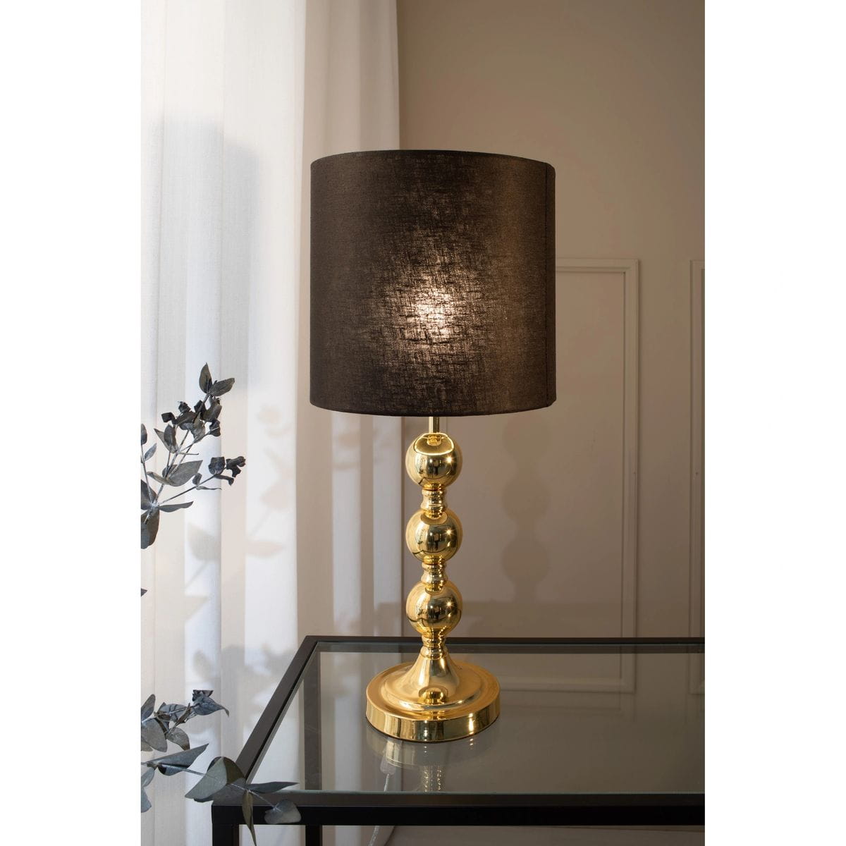 Lampa stołowa Octo z mosiądzu i czarnym abażurem, zbudowana z trzech kulistych elementów, umieszczona na okrągłej podstawie. Ujęcie frontalne, lampa stoi na szklanym stoliku w jasnym wnętrzu, obok zasłon i rośliny.
