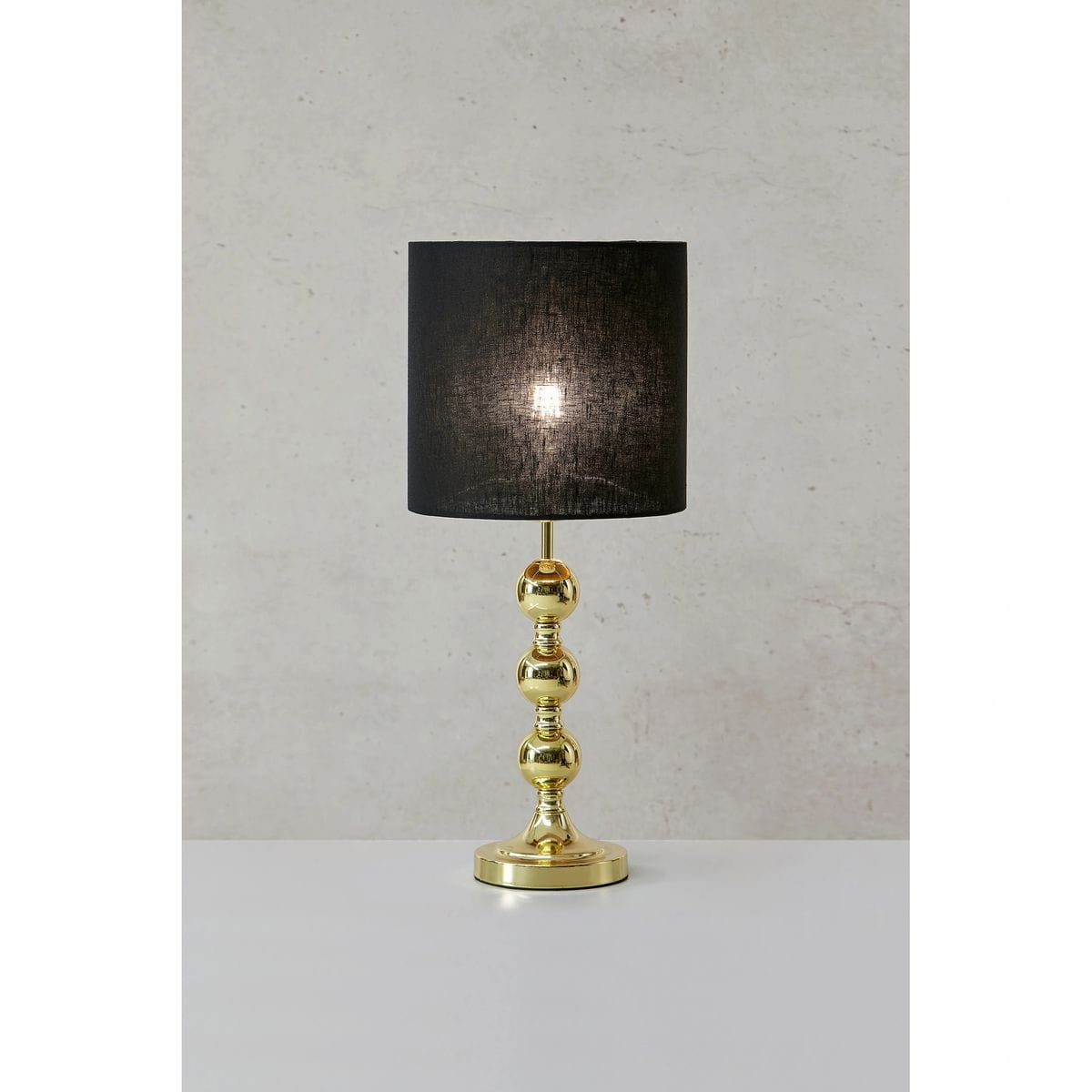 Lampa stołowa Octo z mosiądzu w kolorze złotym, z czarnym abażurem. Ułożona pionowo na płaskiej powierzchni, z widocznymi kulistymi detalami podstawy. Abażur wykonany z tkaniny, emitujący światło. Tło w neutralnym kolorze.