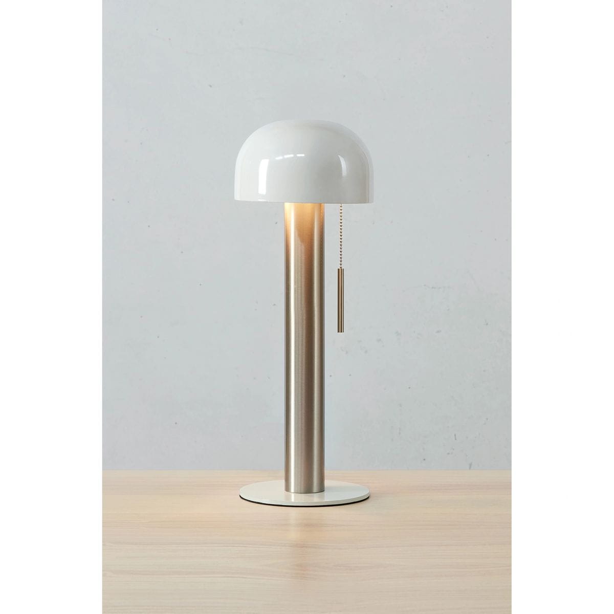 Lampa stołowa Costa z niklowanym, satynowym trzonem i białym kloszem w kształcie półkuli. Widoczny metalowy łańcuszek do włączania/wyłączania. Produkt umieszczony na drewnianym blacie, z neutralnym tłem.