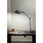 Portland lampa biurkowa z metalowym, chromowanym kloszem i regulowanym ramieniem. Ustawiona na drewnianym stole obok papierowego kubka i owalnego podkładki. Tło w jasnych odcieniach, subtelne oświetlenie.