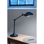 Czarna lampa biurkowa Portland z regulowanym ramieniem, wyposażona w klosz o zaokrąglonym kształcie. Ustawiona na drewnianym biurku, obok okrągłej podkładki i długopisu, w tle widoczny jasny okno.