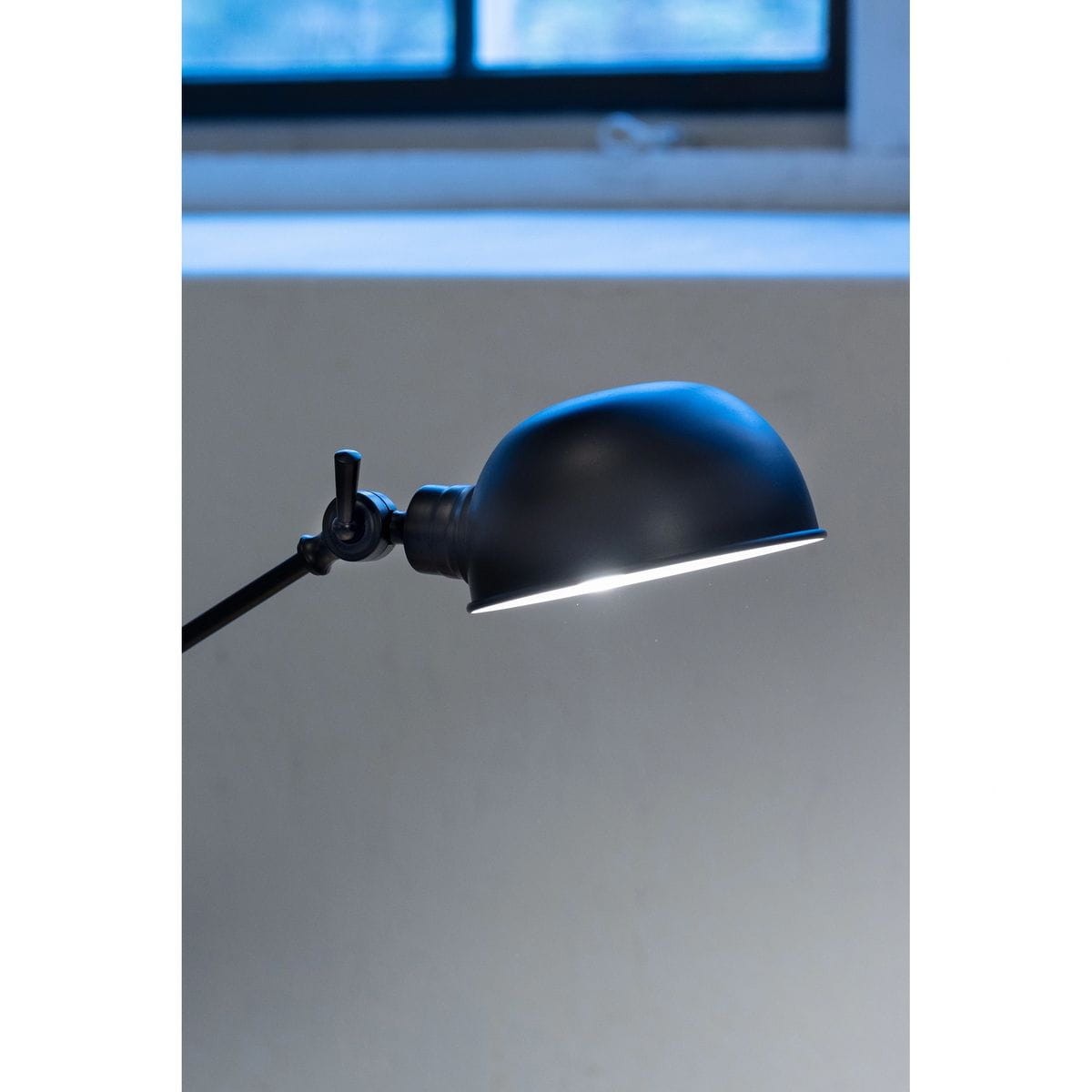 Czarna lampa biurkowa Portland z kloszem w formie półkuli, montowana na regulowanym ramieniu. Widoczny element mocujący. Lampa przeznaczona do żarówki E27, umiejscowiona w pobliżu okna. Tło w odcieniach szarości.