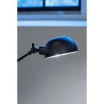 Czarna lampa biurkowa Portland z kloszem w formie półkuli, montowana na regulowanym ramieniu. Widoczny element mocujący. Lampa przeznaczona do żarówki E27, umiejscowiona w pobliżu okna. Tło w odcieniach szarości.