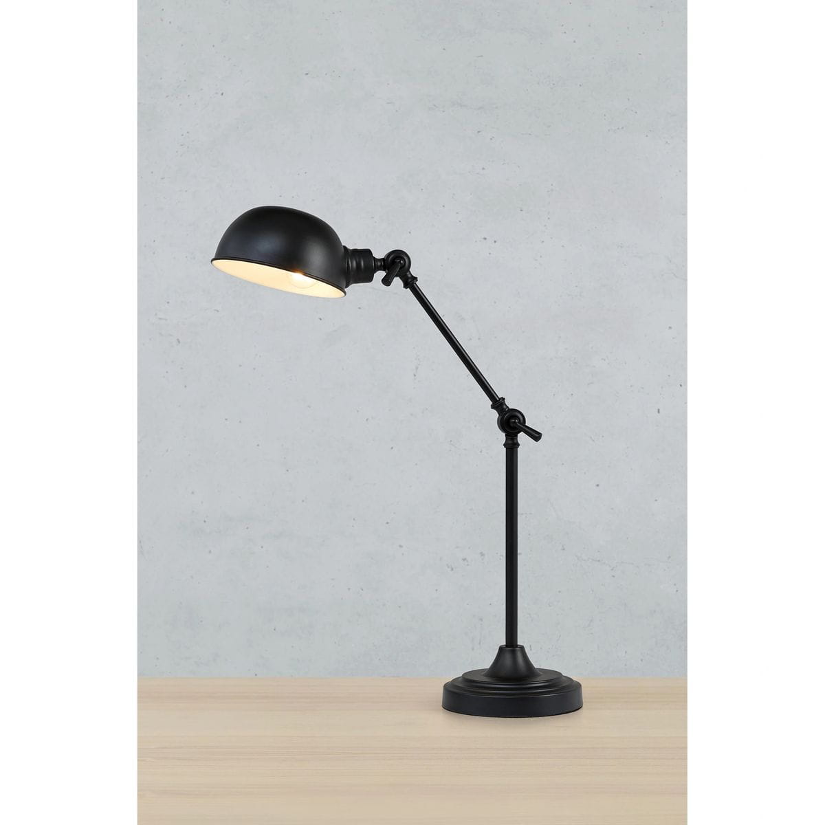 Czarna lampa biurkowa Portland z metalowym kloszem i regulowanym ramieniem. Stojąca na okrągłej podstawie, z widoczną oprawą na żarówkę E27. Ujęcie z boku, na tle szarej ściany, z drewnianym blatem.