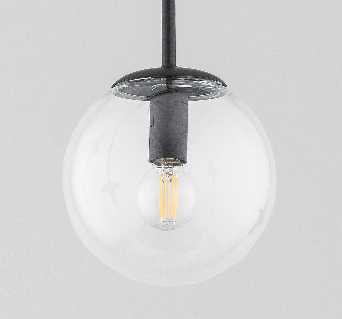 Klosz szklany Sebia kula D150, przezroczysty, w kształcie kuli, zawieszony na czarnym przewodzie. Wewnątrz widoczna żarówka z widocznymi włóknami. Tło jasnoszare, co podkreśla przezroczystość szkła.