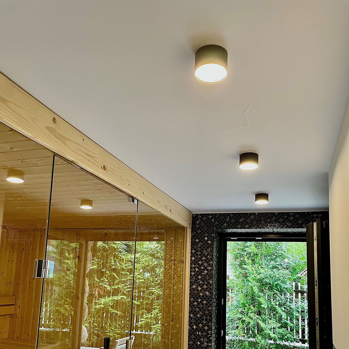 Czarna lampa sufitowa Kendal LED IP54 w kształcie cylindra, zamontowana na białym suficie. Oświetlenie skierowane w dół, w otoczeniu drewnianej ścianki i lustrzanej powierzchni. Widoczna część pokoju z drzwiami prowadzącymi na zewnątrz.