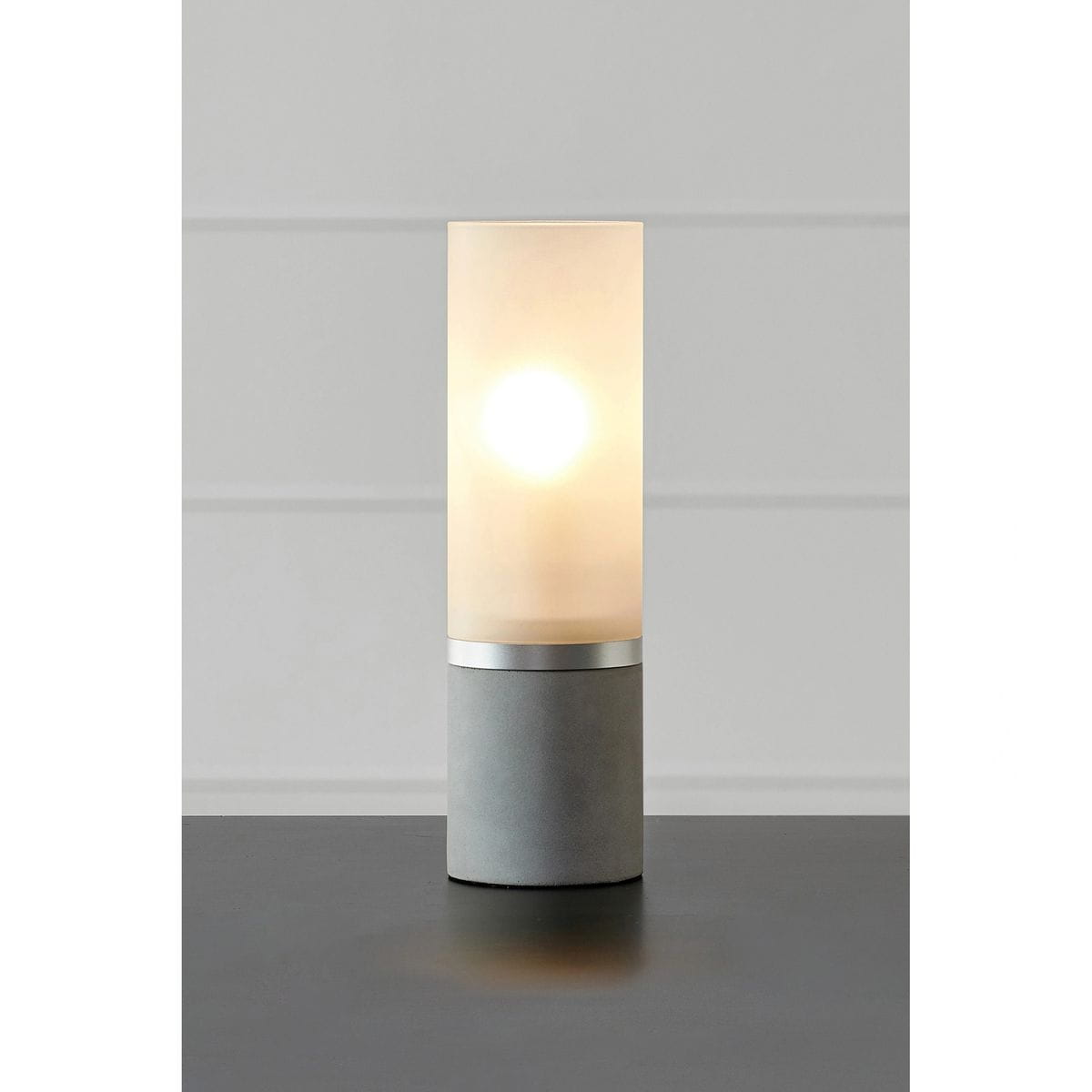 Lampa stołowa Molo 1xE14 w szarym kolorze z cylindrycznym, matowym kloszem i metalowym pierścieniem. Ujęcie frontalne, stojąca na czarnej powierzchni, emitująca ciepłe światło. Idealna do nowoczesnych wnętrz.