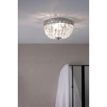 Lampa sufitowa Etienne z chromowanym wykończeniem, z dwiema oprawami E14. Okrągła konstrukcja z przezroczystymi kryształkami, montowana na suficie, emitująca miękkie światło w neutralnym wnętrzu. Widoczny fragment ściany w tle.