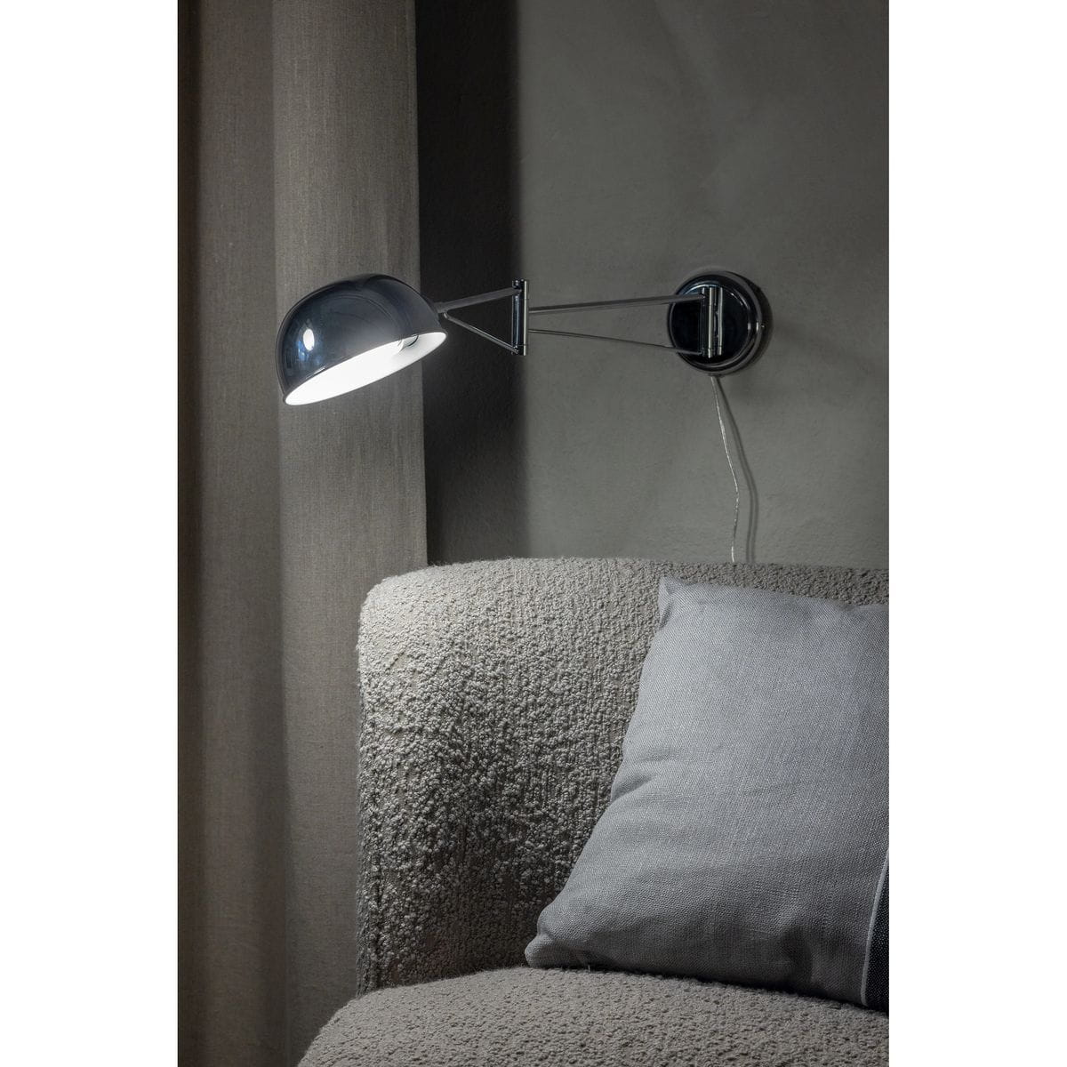Kinkiet Portland 1xE27 w chromowanym wykończeniu, z okrągłą, błyszczącą czaszą. Lampa zamontowana na ścianie obok szarej kanapy z poduszką. Widoczny zintegrowany kabel. Tło w odcieniach szarości, tworzy elegancką aranżację.