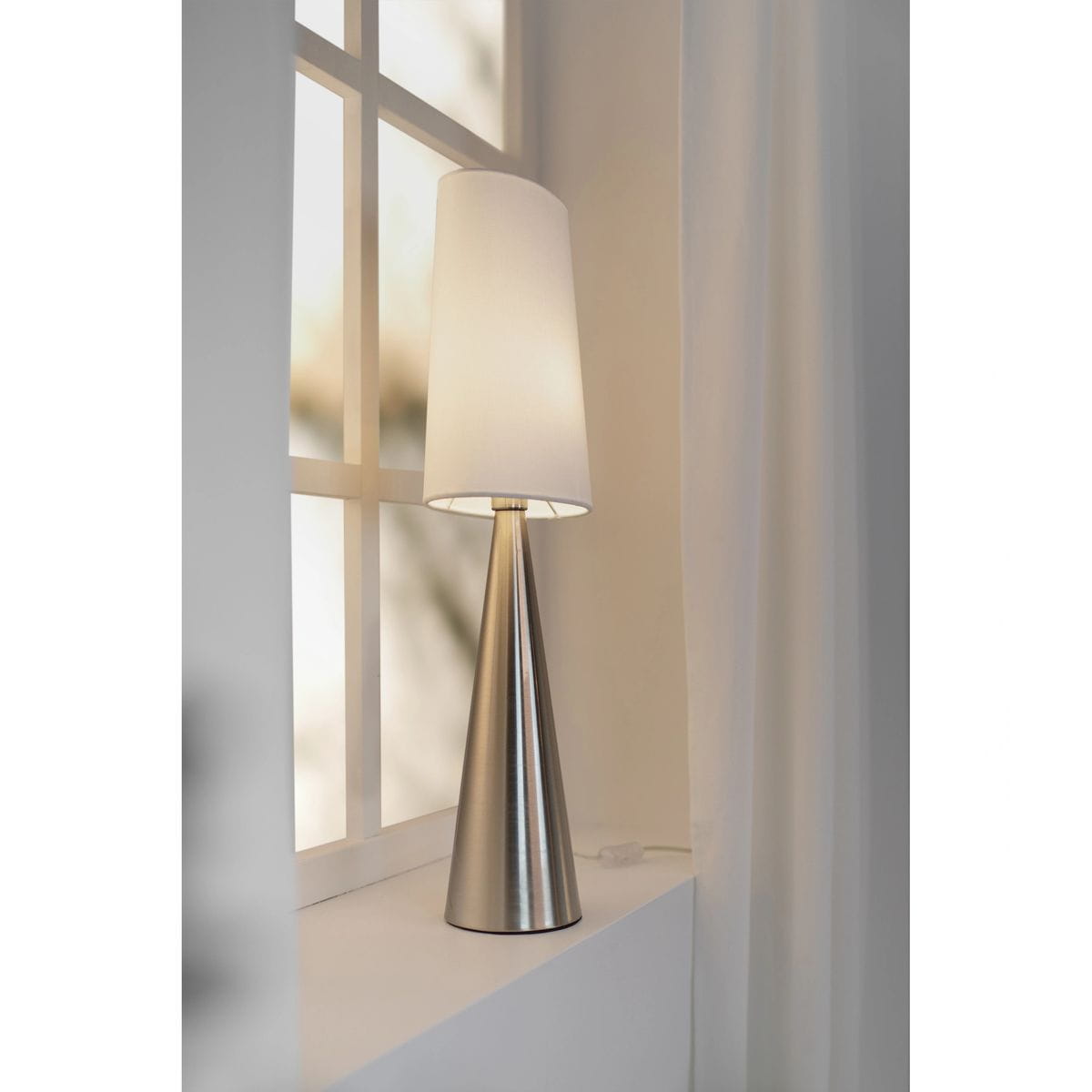 Lampa stołowa Conus z materiału niklowego w satynowym wykończeniu, z białym kloszem w kształcie stożka. Ustawiona na białym parapecie, z tyłu widoczne okno. Nogi lampy stożkowate, zapewniające nowoczesny wygląd.