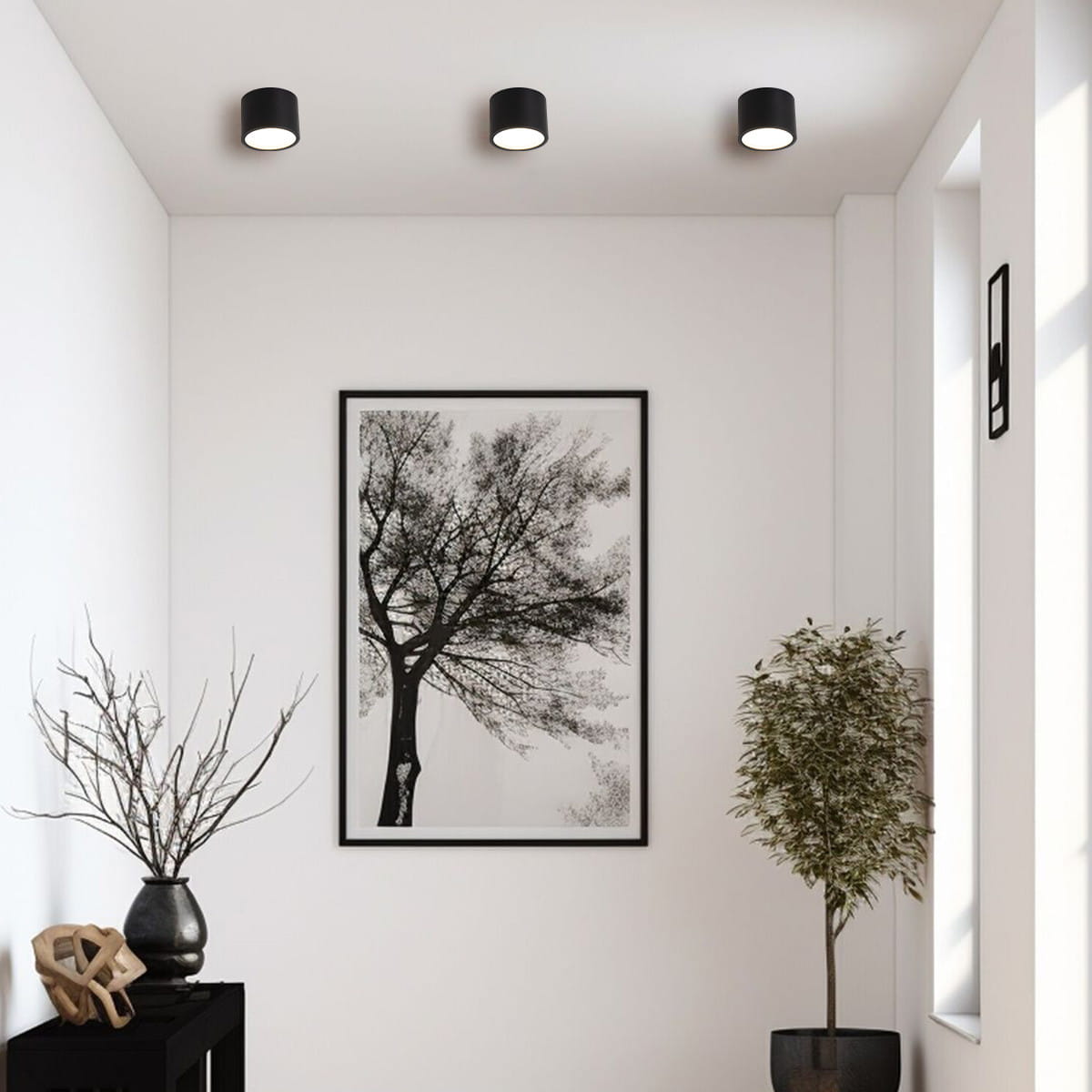 Czarna lampa sufitowa tuba Kendal IP65 z jedną oprawą GX53, zamontowana na suficie w minimalistycznym wnętrzu. Widoczne także drzewo w ramie oraz roślina w doniczce na stoliku. Tło białe, jasne i neutralne.
