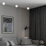 Lampa sufitowa Kendal 1xGX53 w kolorze białym, zamontowana na suficie. Okrągły kształt, matowa powierzchnia. W tle widoczna szara ściana, zasłony z wzorem oraz kanapa z poduszkami.