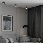 Czarna lampa sufitowa Kendal (spot) 1xGX53 z ruchomą głowicą, zamontowana na białym suficie. W tle widoczna szara sofa z poduszkami oraz ciemne zasłony z wzorem. Obok obraz w ramie z abstrakcyjnym motywem.