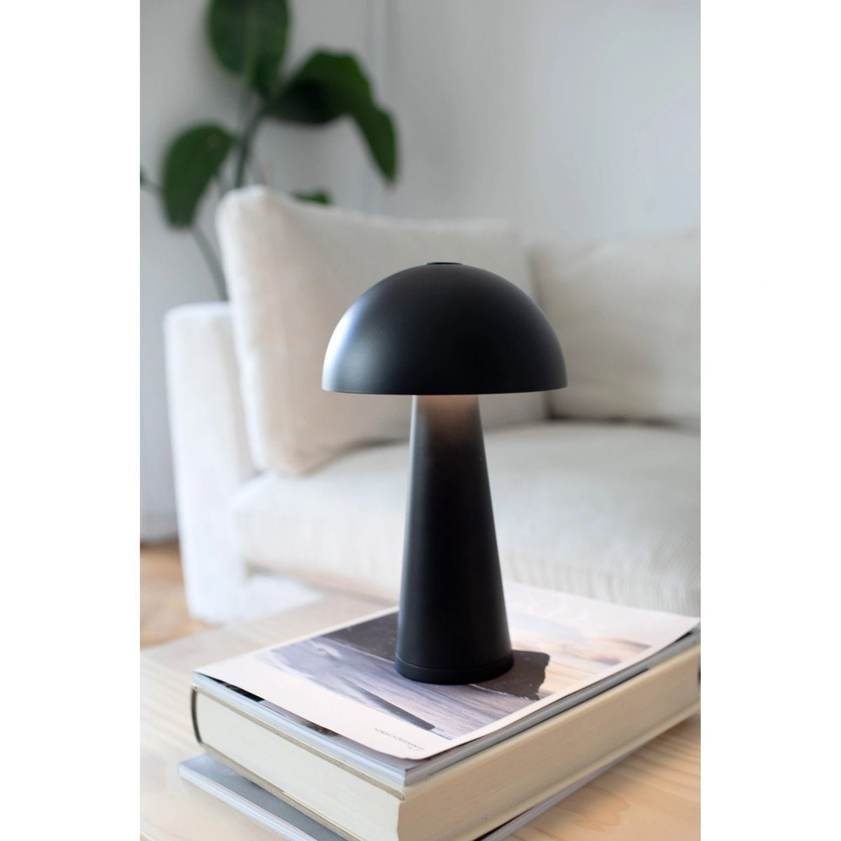 Czarna lampa stołowa Fungi z akumulatorem, o kształcie grzyba, z kloszem w formie półkuli i stożkowatą podstawą. Ustawiona na książkach na stoliku, w tle widoczna kanapa i roślina. Wbudowana dioda LED o mocy 1,5W.