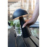 Czarna lampa stołowa akumulatorowa Fungi z jedną żarówką LED 1,5W, trzymana w dłoni. Posiada okrągły klosz i cylindryczną podstawę. Otoczenie: stół z drewnianymi deskami i kwiaty w wazonie.