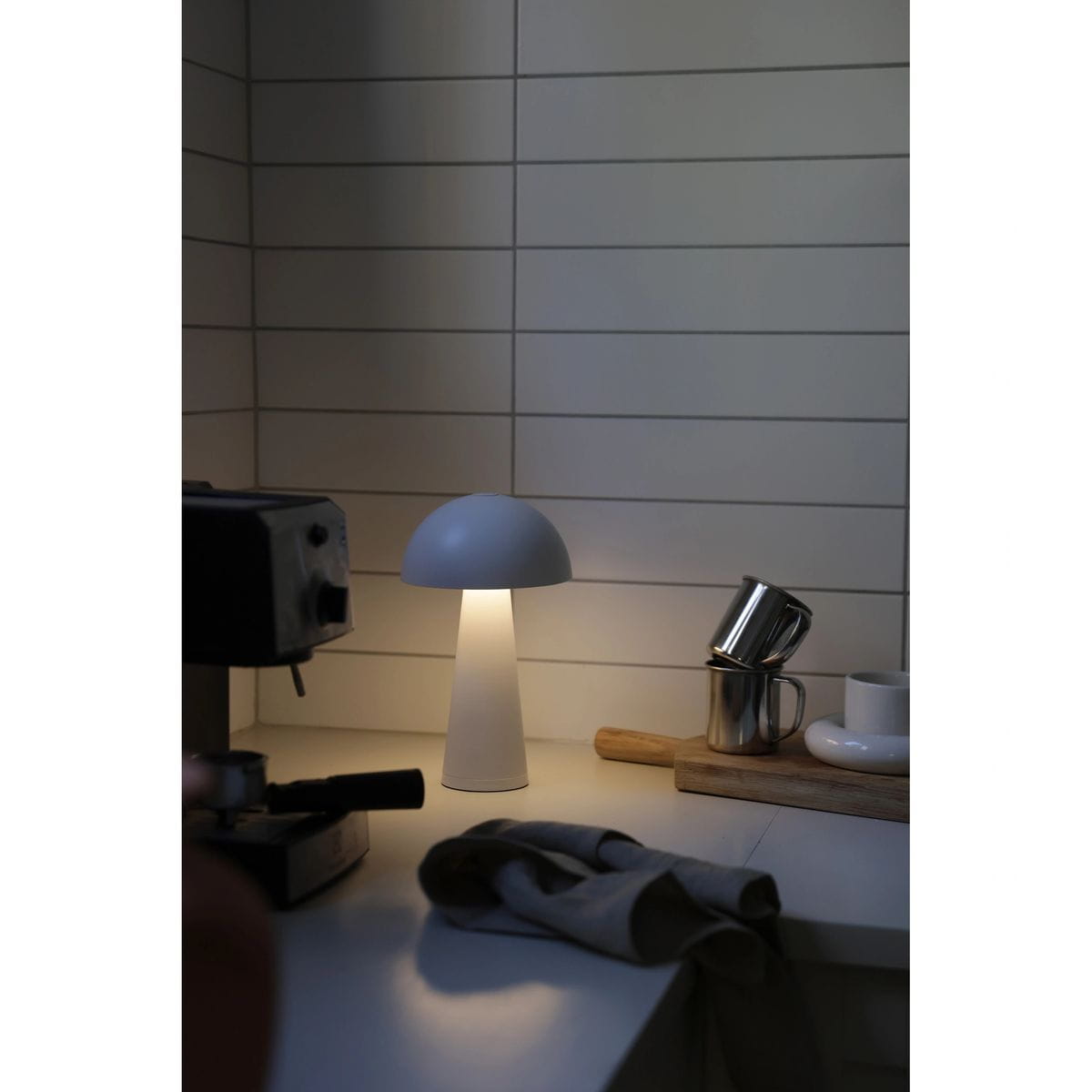 Lampa stołowa Fungi w kolorze białym, z akumulatorem, z kloszem w kształcie grzyba. Ujęcie z boku, stojąca na blacie, w otoczeniu filiżanek i akcesoriów kuchennych. Emituje miękkie światło, tworząc przytulną atmosferę.