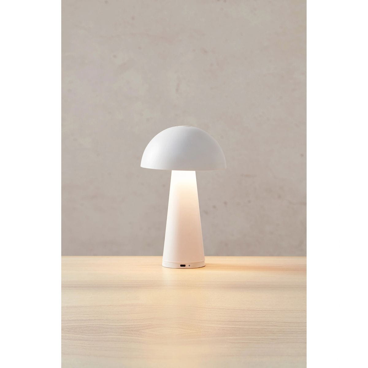 Biała lampa stołowa akumulatorowa Fungi z kloszem w kształcie półkuli i podstawą stożkową. Ujęcie frontalne, lampa emituje ciepłe światło, umieszczona na drewnianym stole, z widocznym przyciskiem na podstawie.