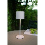 Lampa stołowa Filo, akumulatorowa, biała, z cylindrycznym kloszem, na cienkiej nodze i okrągłej podstawie. Zbliżenie z boku, na stole, w otoczeniu zieleni, z delikatnym światłem LED.