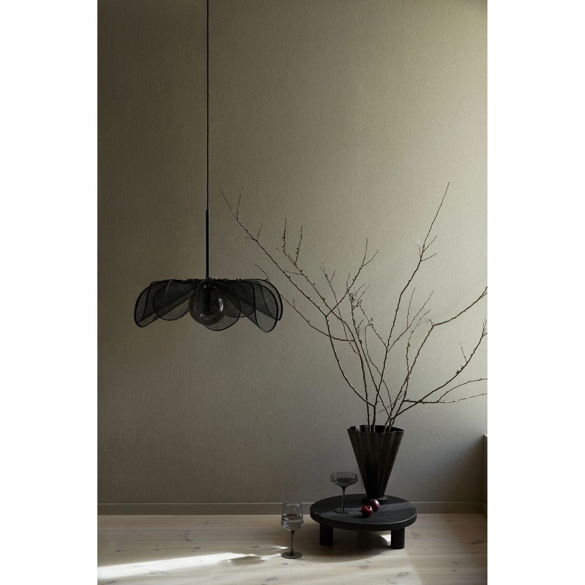 Lampa wisząca Styrka w kolorze czarnym z dymnym kloszem, zawieszona na cienkim przewodzie. Kształt klosza przypomina kwiat, a lampa jest umieszczona w minimalistycznym otoczeniu z drewnianą podłogą i dekoracyjnymi elementami.