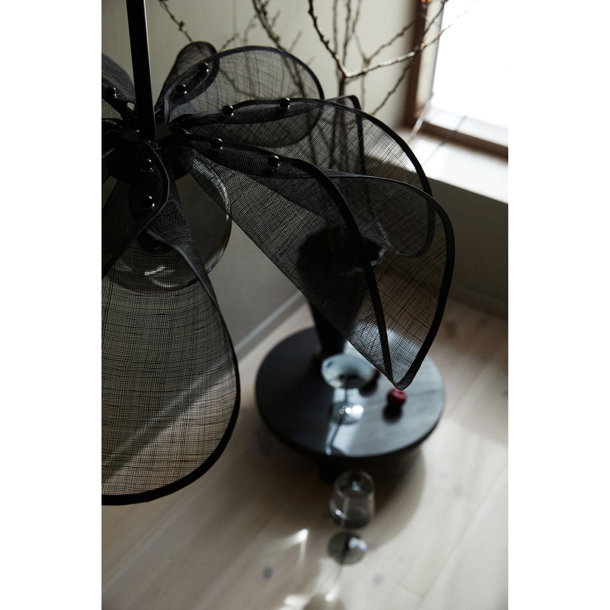 Lampa wisząca Styrka 1xE27 w kolorze czarnym z dymnym kloszem, z charakterystycznymi, szerokimi, półprzezroczystymi elementami. Ujęcie z góry, ukazujące detale klosza oraz okrągłą podstawę. W tle widać drewnianą podłogę i szklane akcesoria.