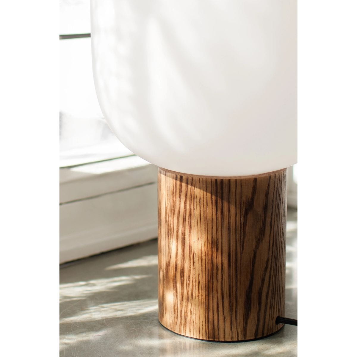 Lampa stołowa Skene z ciemnego drewna, z okrągłą podstawą i białym, matowym kloszem. Produkt widoczny z boku, z detalem podstawy, ukazującym naturalną fakturę drewna. Kabel zasilający widoczny obok.