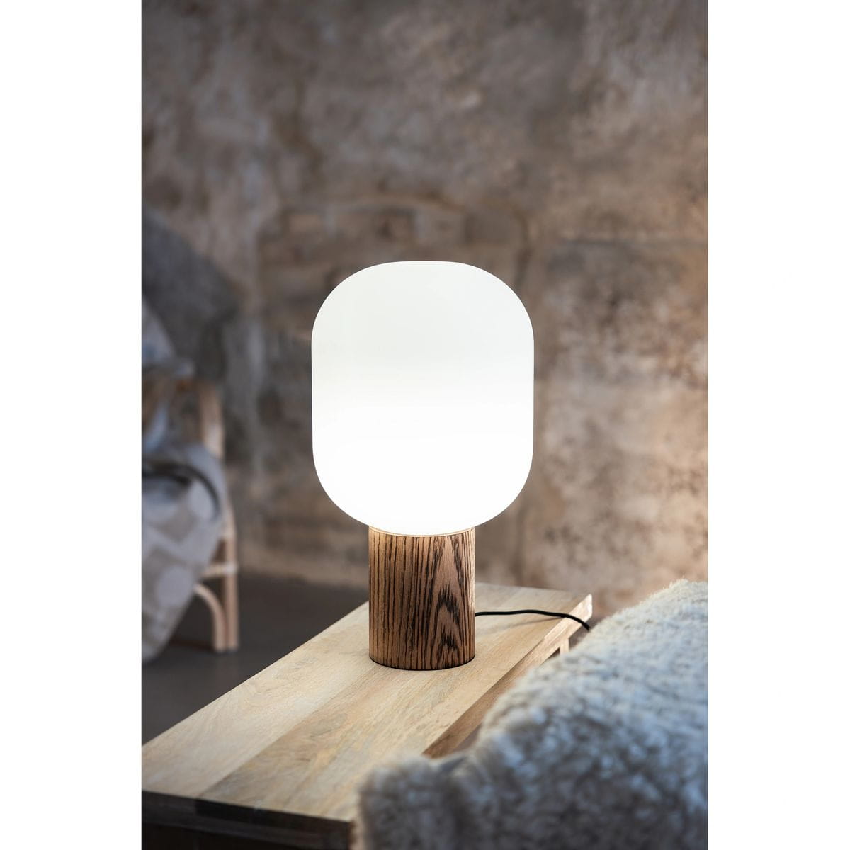 Lampa stołowa Skene 1xE27 z ciemnego drewna, z okrągłą, matową, białą kloszem. Produkt stoi na drewnianej powierzchni, obok znajduje się miękka, jasna tkanina. Klosz ma zaokrąglony kształt, a drewno ma wyraźne słoje.