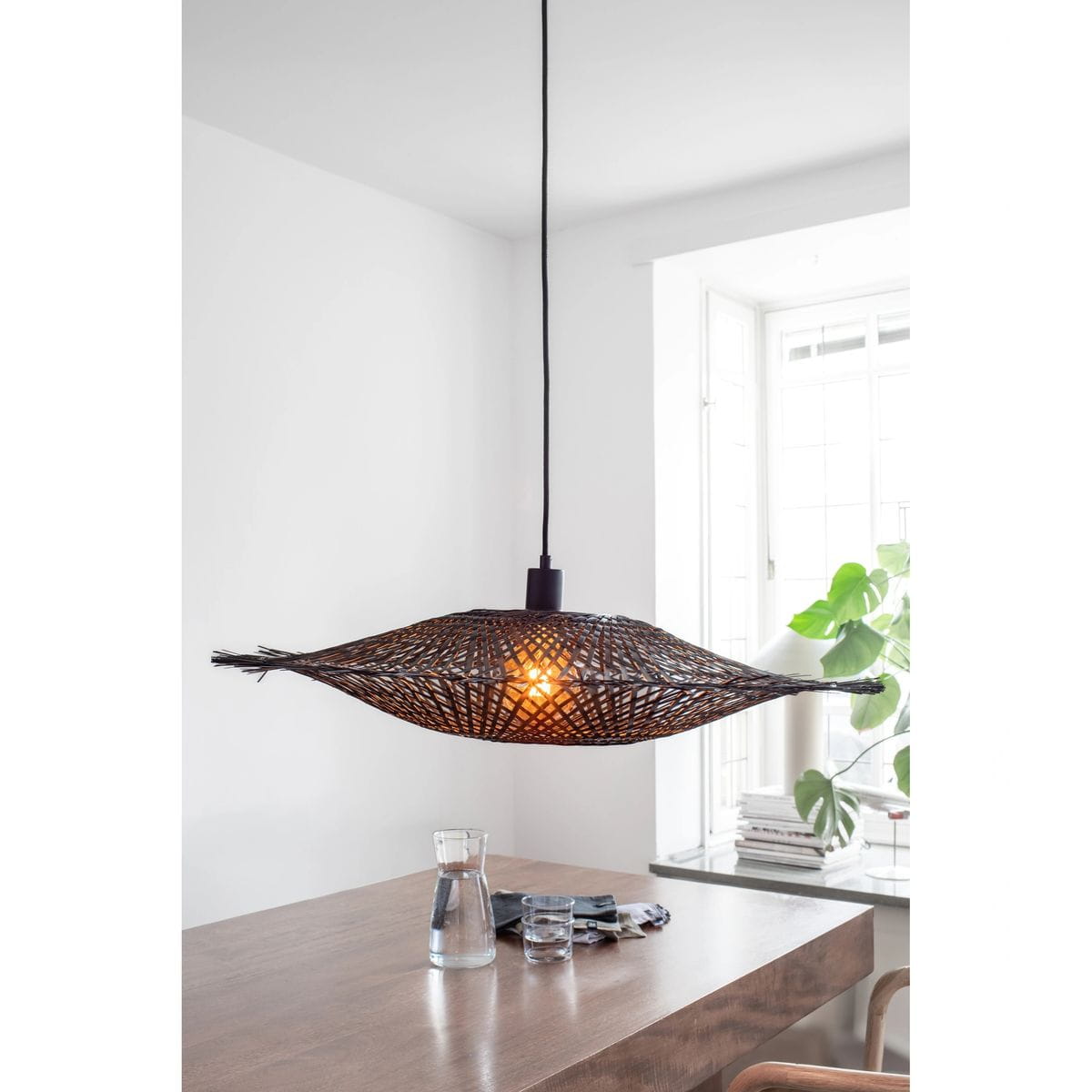Czarna lampa wisząca Kumo z jedną oprawą E27, o organicznym, plecionym kształcie przypominającym liść. Zawieszona na czarnym przewodzie, emituje ciepłe światło, tworząc przytulną atmosferę. Widoczna w nowoczesnym wnętrzu z drewnianym stołem i roślinami.